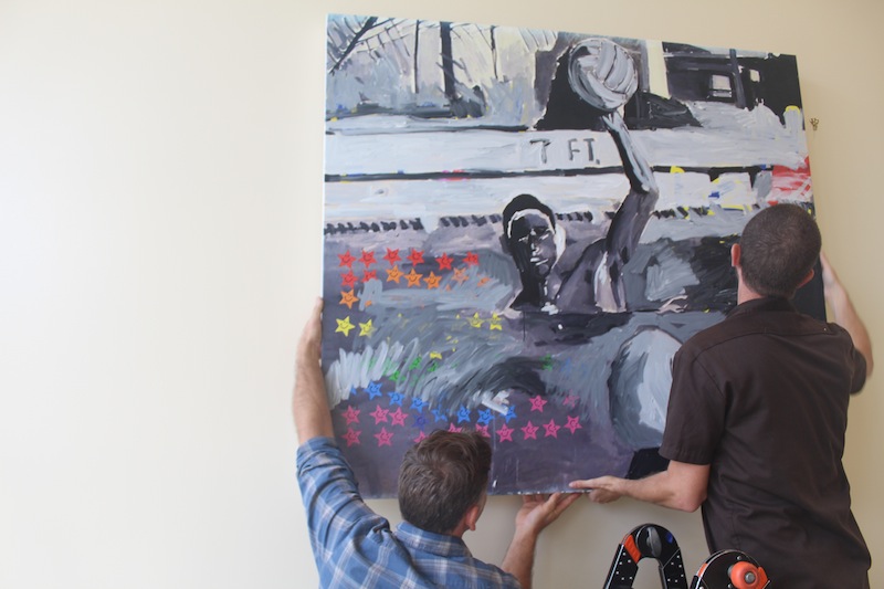 Two helpers hang up Franco's painting of a water polo player in 1993. Over 10 of Franco's paintings that are inspired from yearbook photos are spread throughout the Media Arts center. Photo by Adele Bloch.