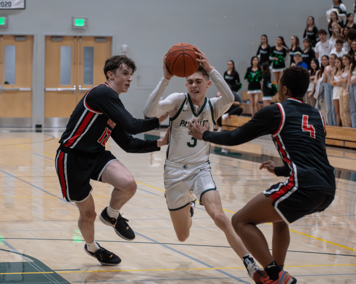 Palo Alto High School Viking junior point guard Gavin Haase attempts a layup while two Henry M. Gunn High School Titans try to block the shot Saturday night during the second crosstown rivalry match on Saturday night. The Vikings narrowly lost to the Titans, 63-65, after the game extended to overtime following a fourth quarter comeback by the Vikings. According to Viking senior shooting guard Riley Yuen, although the Vikings gained momentum from the comeback, they were unable to secure a win in the closing moments of the game. “We felt really confident going overtime,” Yuen said. “We knew we would have to lock in on defense. We had our chances on offense, but we missed too many open hoops.” (Photo: Maxwell Zhang)
