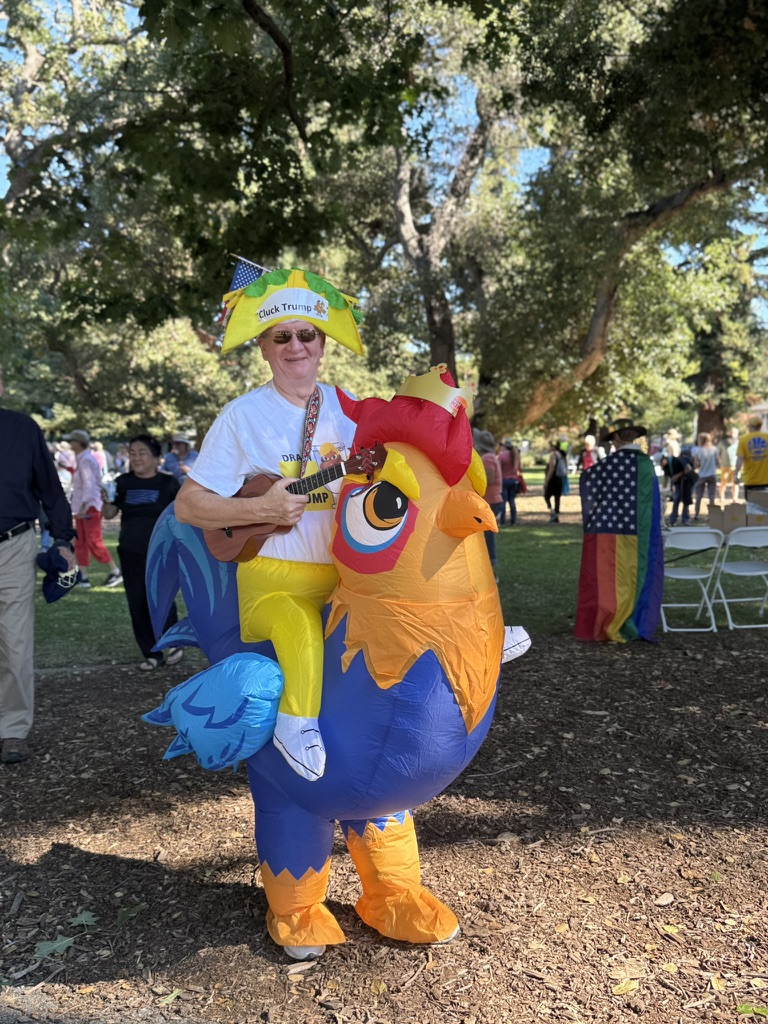 A protester in a chicken suit wearing a taco hat with the words “Cluck Trump” at the “No Kings” Democracy Fair in Palo Alto on Saturday. Protestor Mary Chan said she and her husband are horrified at the state of science and medicine in America. “What he [Trump] is doing by defunding scientific research is destroying our academic medical centers, and he’s [Trump] bankrupting rural medical centers,” Chan said. “We will have lasting negative impacts on American health for decades.”